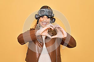 Young beautiful blonde aviator woman wearing vintage pilot helmet whit glasses and jacket smiling in love doing heart symbol shape