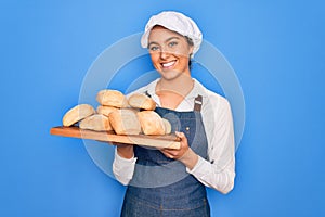 Young beautiful blonde baker woman with blue eyes holding tray with homemade fresh bread with a happy face standing and smiling