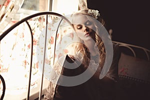 Young beautiful blonde in the bed at morning time near window under Sun rays with flower in the hair. Tender romantic mood