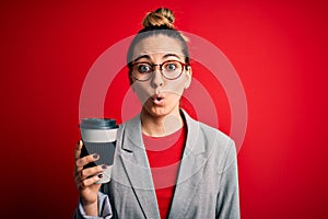 Young beautiful blonde businesswoman with blue eyes wearing glasses drinking cup of coffe scared in shock with a surprise face,