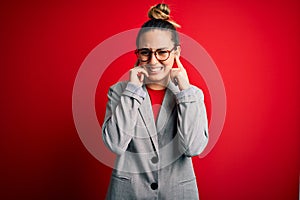 Young beautiful blonde businesswoman with blue eyes wearing glasses and jacket covering ears with fingers with annoyed expression