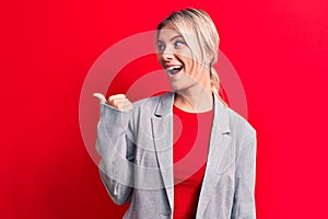 Young beautiful blonde businesswoman wearing elegant jacket over isolated red background pointing thumb up to the side smiling