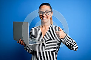 Young beautiful blonde businesswoman wearing glasses using laptop over blue background happy with big smile doing ok sign, thumb