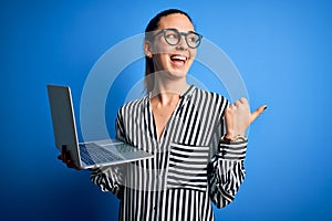 Young beautiful blonde businesswoman wearing glasses using laptop over blue background pointing and showing with thumb up to the