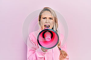 Young beautiful blonde caucasian woman shouthing and screaming through megaphone over isolated pink background