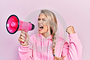 Young beautiful blonde caucasian woman shouthing and screaming through megaphone over isolated pink background