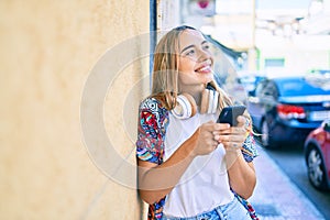 Young beautiful blonde caucasian woman smiling happy outdoors on a sunny day wearing headphones and using smartphone
