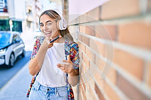 Young beautiful blonde caucasian woman smiling happy outdoors on a sunny day wearing headphones and using smartphone leaning on a