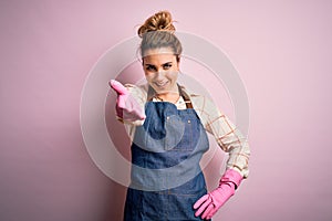 Young beautiful blonde cleaner woman doing housework wearing arpon and gloves smiling friendly offering handshake as greeting and