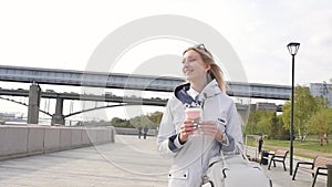 Young beautiful blonde drinks her coffee and enjoys a walk on the embankment of the river. Slow motion