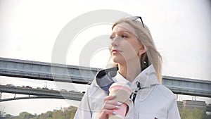 Young beautiful blonde drinks her coffee and enjoys a walk on the embankment of the river. Slow motion