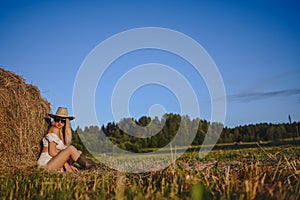 Young beautiful blonde farmer woman in white outfit, sunglasses cowboy hat and boots posing at sunset in field with