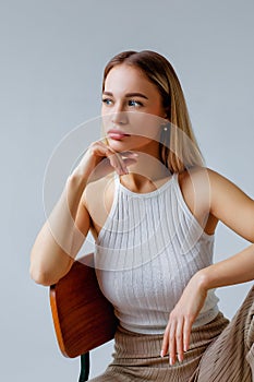 A young beautiful blonde in a fashionable white T-shirt and beige knitted trousers is sitting on a chair, looking away on a gray