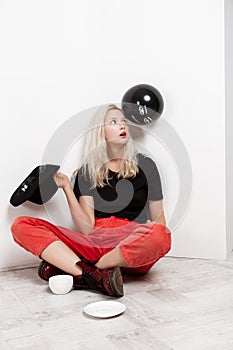 Young beautiful blonde girl with black baloon in hat sitting on floor drinking coffee over white wall.