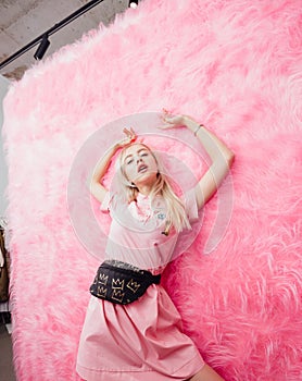 Young beautiful blonde girl dressed in fashion pink dress poses on the background of pink fur wall in the show room