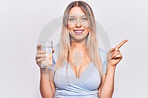 Young beautiful blonde girl drinking glass of water smiling happy pointing with hand and finger to the side