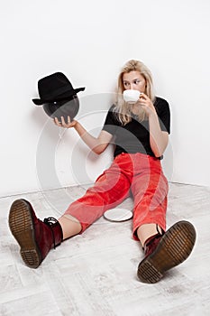 Young beautiful blonde girl holding black baloon in hat sitting on floor drinking coffee over white wall.