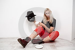 Young beautiful blonde girl holding black baloon in hat sitting on floor drinking coffee over white wall.