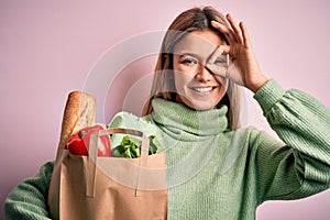 Young beautiful blonde girl holding fresh groceries paper bag over pink isolated background with happy face smiling doing ok sign