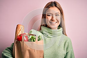 Young beautiful blonde girl holding fresh groceries paper bag over pink isolated background with a happy face standing and smiling