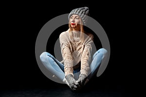 Young beautiful blonde girl in knitted gloves and hat in gray, blue jeans, beige sweater sits on the floor in a butterfly pose