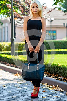 Young beautiful blonde girl posing on the background of the urban landscape. lady in a black dress and red shoes.