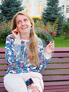 Young beautiful blonde girl sitting on a bench in the park with a smartphone smiling, speaking on the phone, listening to music.