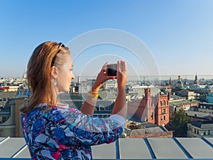 Young beautiful blonde girl taking pictures of the city with a smartphone on a rooftop in Moscow, Russia.