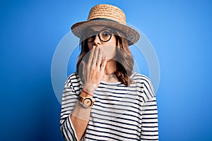 Young beautiful blonde girl wearing glasses and hat over blue  background cover mouth with hand shocked with shame for