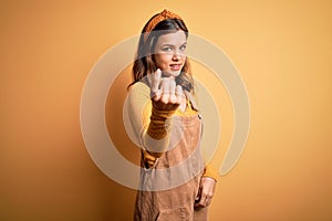 Young beautiful blonde girl wearing overall standing over yellow isolated background Beckoning come here gesture with hand