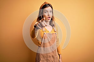 Young beautiful blonde girl wearing overall standing over yellow isolated background pointing displeased and frustrated to the