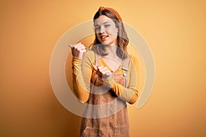 Young beautiful blonde girl wearing overall standing over yellow isolated background Pointing to the back behind with hand and