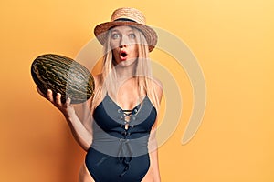 Young beautiful blonde girl wearing swimwear and summer hat holding melon scared and amazed with open mouth for surprise,