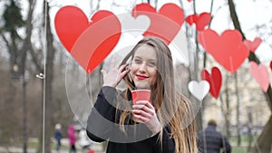 Young beautiful blonde hipster woman posing on the autumn park decorated with red paper hearts background with cup of
