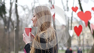 Young beautiful blonde hipster woman posing on the autumn park decorated with red paper hearts background with cup of