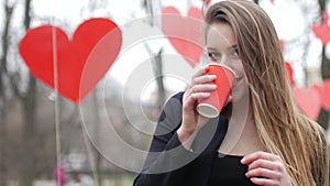 Young beautiful blonde hipster woman posing on the autumn park decorated with red paper hearts background with cup of