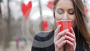 Young beautiful blonde hipster woman posing on the autumn park decorated with red paper hearts background drinking cup