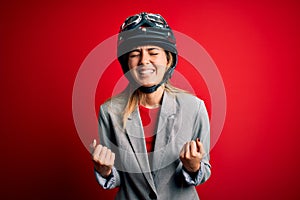 Young beautiful blonde motorcyclist woman wearing motorcycle helmet over red background very happy and excited doing winner