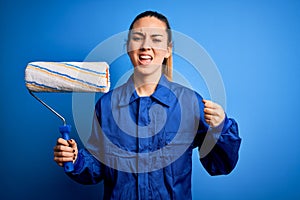 Young beautiful blonde painter woman with blue eyes painting wearing uniform using roller annoyed and frustrated shouting with