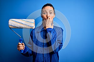 Young beautiful blonde painter woman with blue eyes painting wearing uniform using roller cover mouth with hand shocked with shame