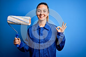 Young beautiful blonde painter woman with blue eyes painting wearing uniform using roller doing ok sign with fingers, excellent