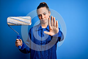 Young beautiful blonde painter woman with blue eyes painting wearing uniform using roller with open hand doing stop sign with