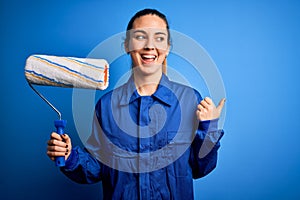 Young beautiful blonde painter woman with blue eyes painting wearing uniform using roller pointing and showing with thumb up to