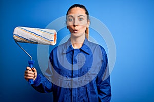 Young beautiful blonde painter woman with blue eyes painting wearing uniform using roller scared in shock with a surprise face,