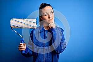 Young beautiful blonde painter woman with blue eyes painting wearing uniform using roller serious face thinking about question,