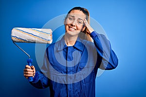 Young beautiful blonde painter woman with blue eyes painting wearing uniform using roller stressed with hand on head, shocked with