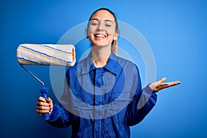 Young beautiful blonde painter woman with blue eyes painting wearing uniform using roller very happy and excited, winner