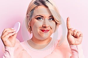 Young beautiful blonde plus size woman applying makeup using sponge over pink background smiling with an idea or question pointing