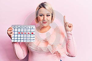Young beautiful blonde plus size woman controlling menstruation holding period calendar smiling with an idea or question pointing