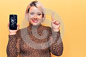 Young beautiful blonde plus size woman holding broken smartphone showing cracked screen smiling with an idea or question pointing